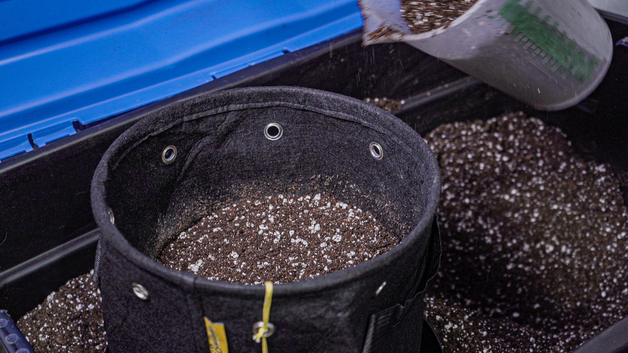 Close-up image of a black fabric BudPots container being filled with soil mix from a gray scooping trowel. The pot is positioned inside a blue storage tub, capturing the motion of soil particles falling into the pot, highlighted by the pot's metal ventilation eyelets.