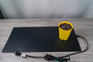 A yellow BudCup filled with soil placed on a black heating mat with an attached electrical cord, set on a grey wood-textured surface against a soft white background.