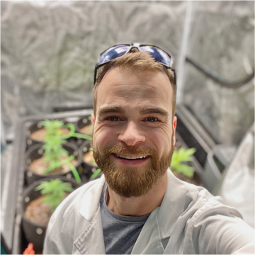 BudTrainer team member in a white lab coat smiling in an indoor cannabis grow tent with healthy plants.
