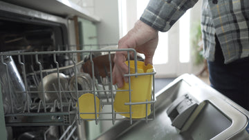 Image of a person's hand placing a yellow BudCups container into a dishwasher rack, among other kitchen items. The person is wearing a gray plaid shirt. The setting suggests the ease of cleaning and maintaining the BudCups container in a typical home environment.
