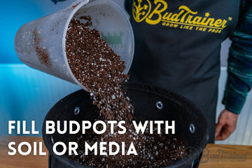 Close-up image of a black fabric BudPots container being filled with soil mix from a gray scooping trowel. The pot is positioned inside a blue storage tub, capturing the motion of soil particles falling into the pot, highlighted by the pot's metal ventilation eyelets.