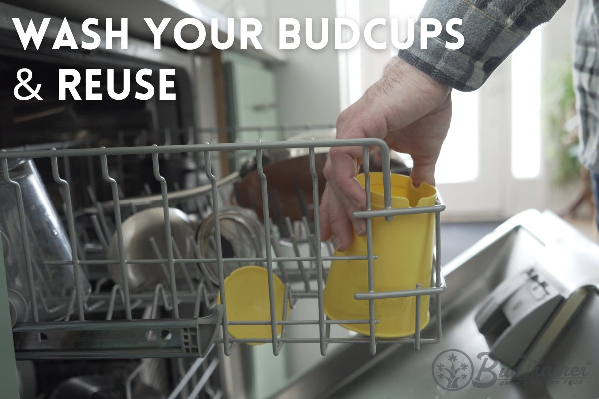 Image of a person's hand placing a yellow BudCups container into a dishwasher rack, among other kitchen items. The person is wearing a gray plaid shirt. The setting suggests the ease of cleaning and maintaining the BudCups container in a typical home environment.